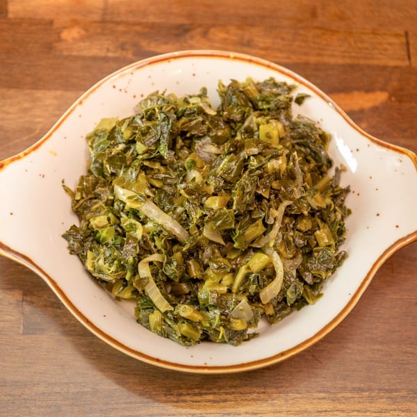 Cooked green leafy vegetables with sliced onions served in a white bowl on a wooden table.