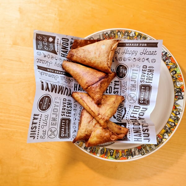 Several golden-brown samosas are served on a white plate with a decorative rim and branded paper.