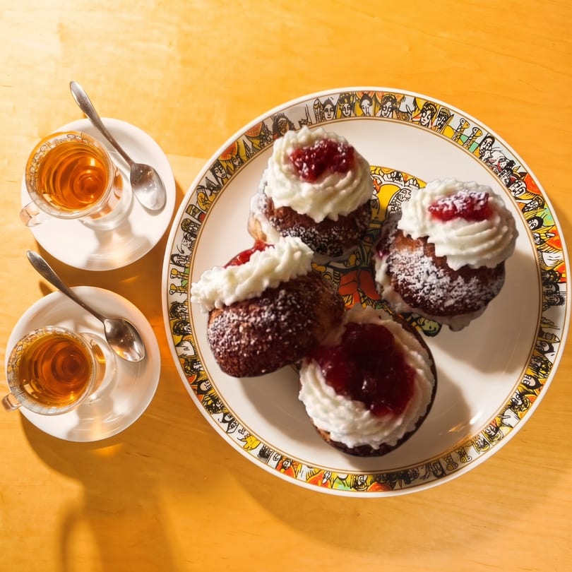 Four pastries with whipped cream and jam on a decorative plate, with two cups of tea on a wooden table.