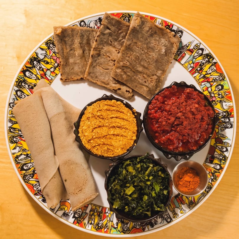 A colorful plate with injera bread, three bowls of different Ethiopian dishes, and a small cup of spice.